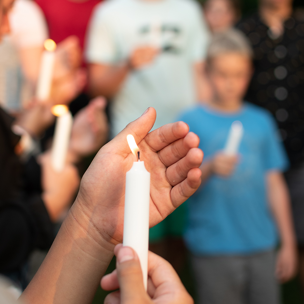 a hand holding a candle stick shielding it from the wind, in the background there are several other students also holding candles