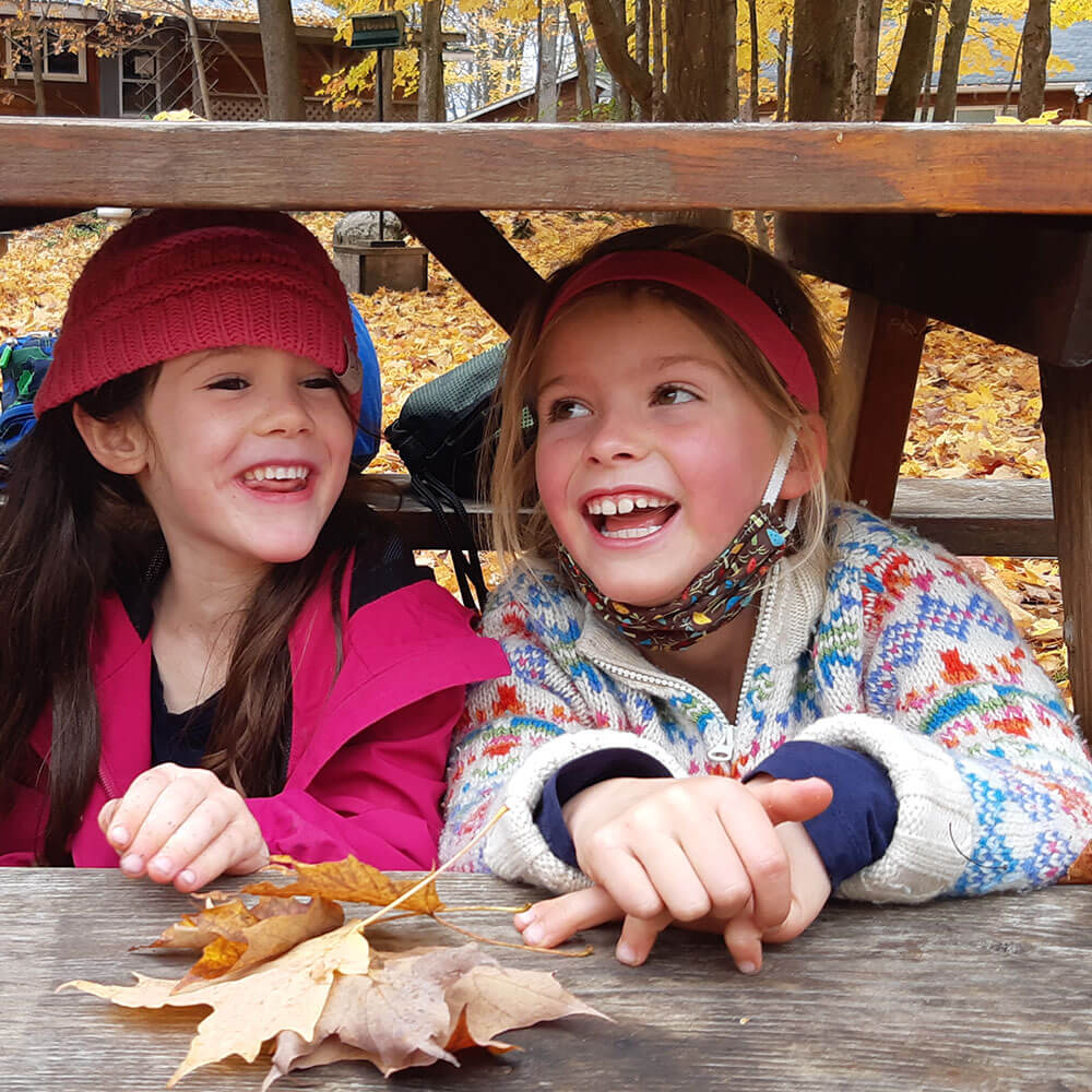 Two smiling young kids under a picnic bench with fall leaves